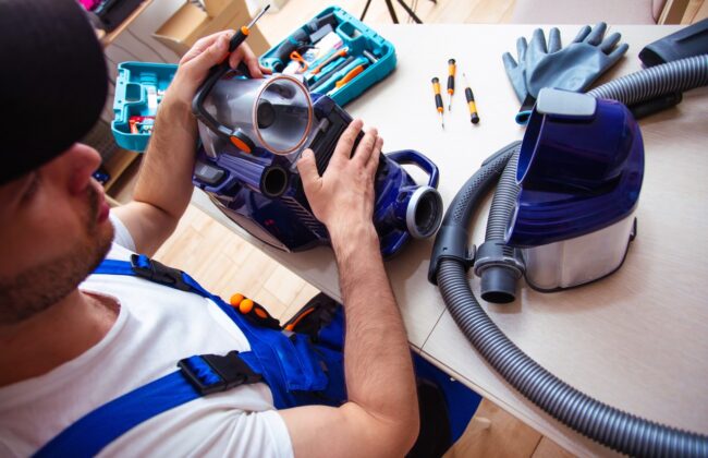 Handsome Young Serviceman Is Repairing Modern Vacuum Cleaner In His Office